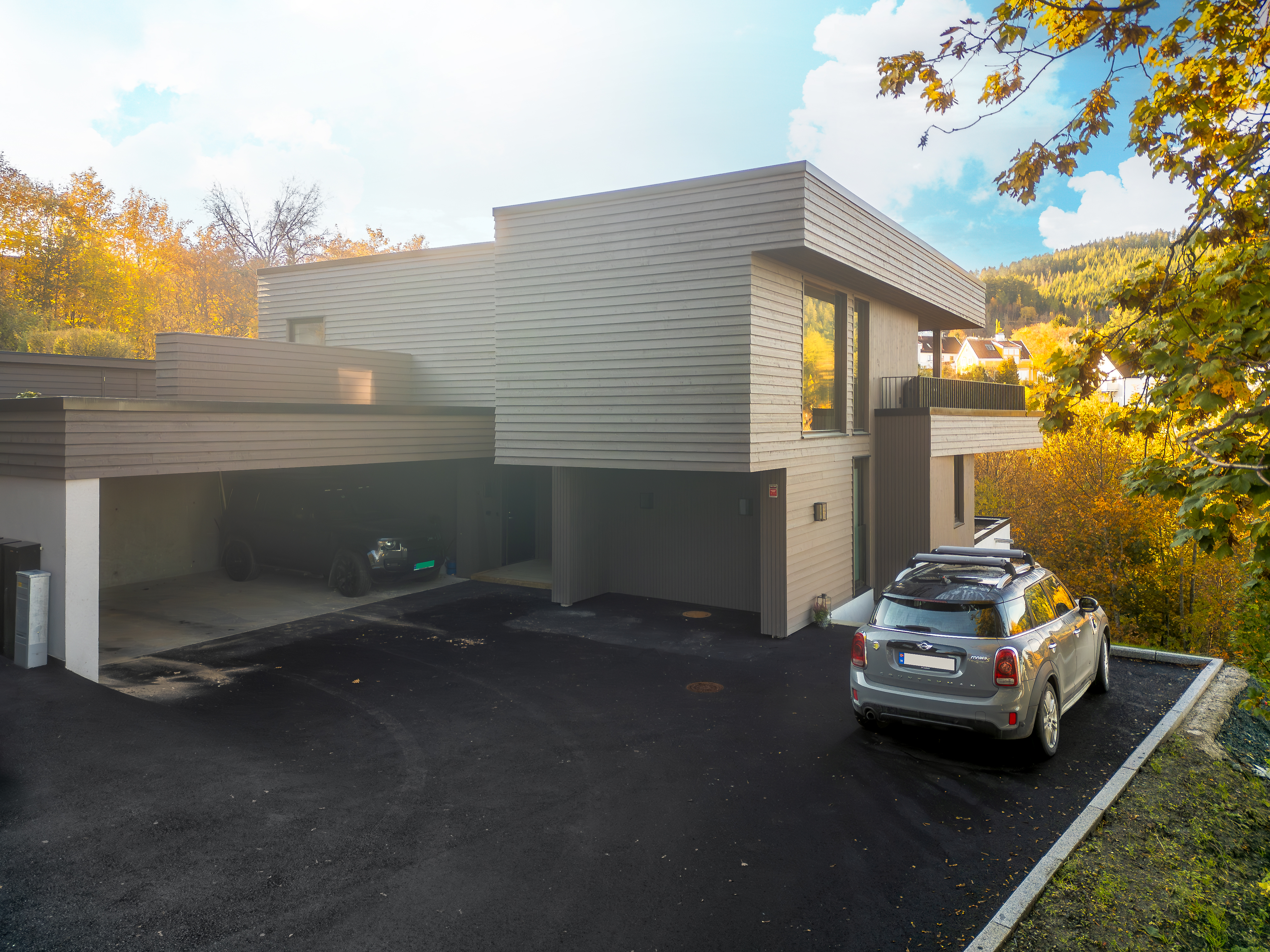 Moderne hus med grå kledning, carport og parkert bil, omgitt av høstløv og fjell.