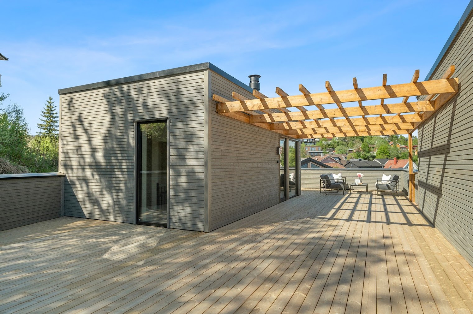 Moderne tilbygg i grått panel med tredekk, pergola og utsikt over boligområde